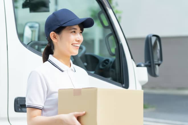 白いポロシャツと紺色のキャップを着用し、笑顔で段ボール箱を抱える女性配送スタッフ