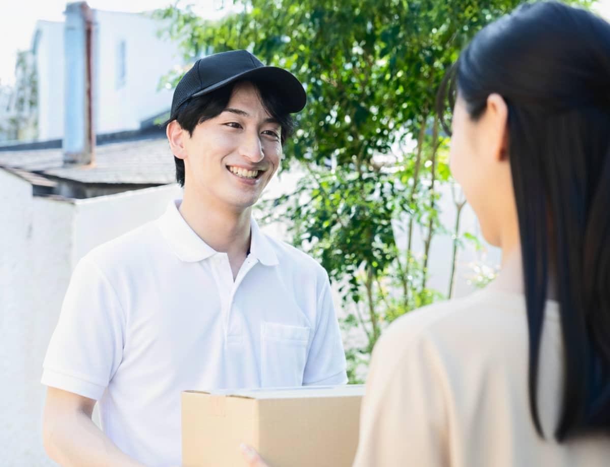 笑顔で段ボール箱を女性に手渡す、白いポロシャツと黒いキャップを被った配達員
