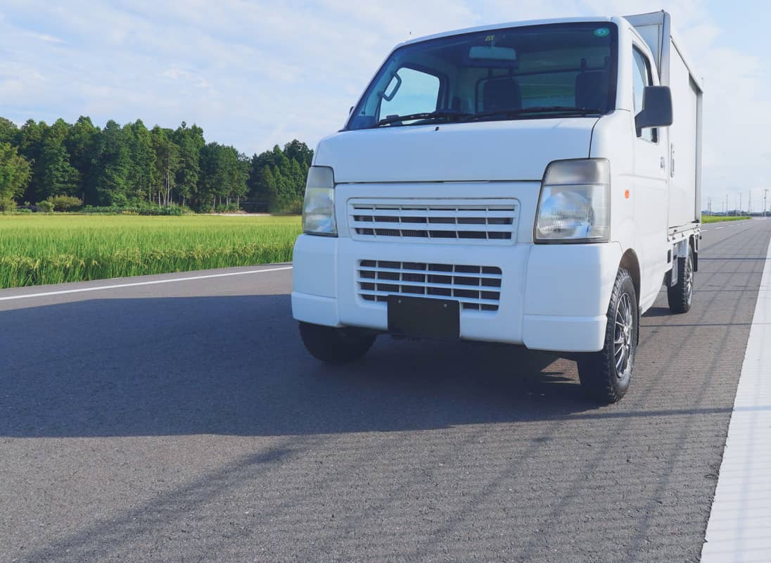 郊外の道路に停車した白い軽貨物トラック。背景には稲が育つ田園と青空が広がる。