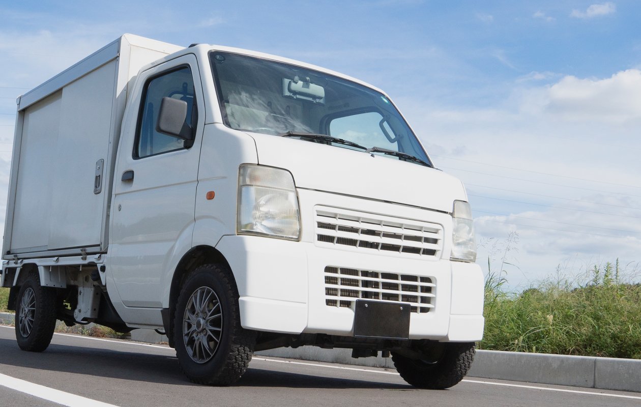 晴天の青空の下、アスファルトの道路を走行する白い軽トラックの斜め前方からの外観。荷台部分はアルミパネルの箱型になっており、配送や運送業務を想起させる清潔感のある商用車の写真。
